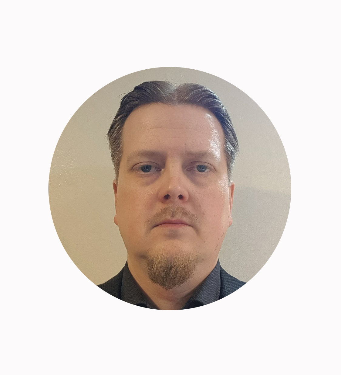 Mies, jolla on lyhyet, liukuhihnaiset ruskeat hiukset, vaalea iho ja pukinparta ja jolla on tumma kauluspaita, katsoo suoraan kameraan vaaleaa taustaa vasten.
