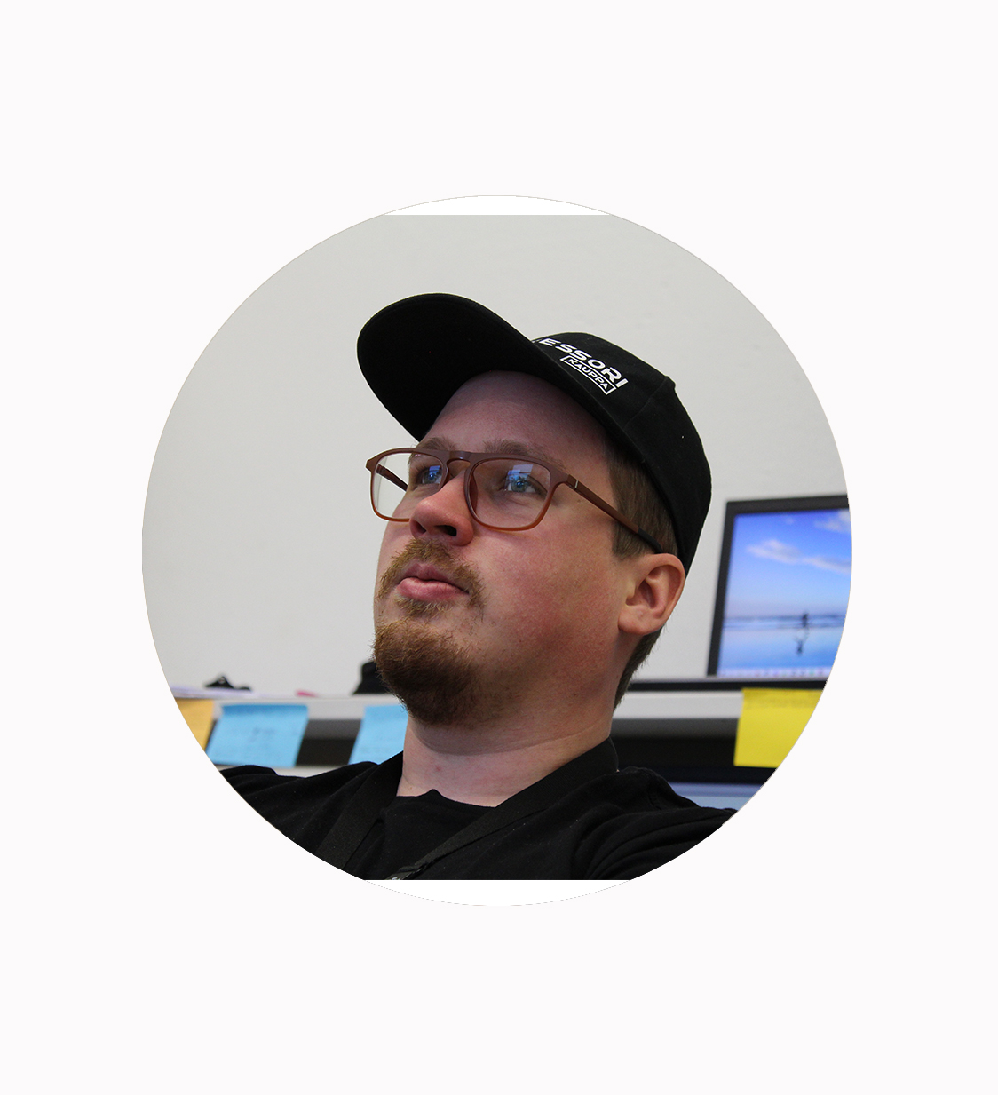 Silmälasipäinen, parrakkaa ja mustaa lippalakkia kantava henkilö katsoo hieman ylöspäin. Taustalla on tietokonemonitori ja useita muistilappuja seinällä. Kuva on rajattu pyöreään kehykseen.